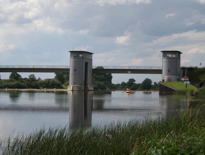 Zwei Türme eines Wehrs reflektieren sich im stillen Wasser, flankiert von grünem Ufergras.