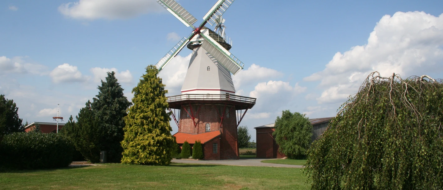 Historische Windmühle umgeben von grüner Wiese und Bäumen, blauer Himmel mit weißen Wolken im Hintergrund.