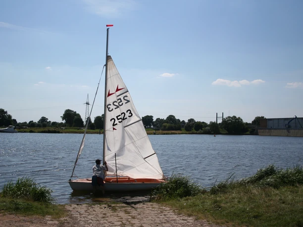 Ein Segelboot mit weißem Segel liegt am Ufer eines ruhigen Flusses, klare Sommertagstimmung.