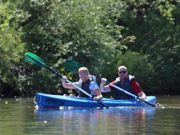 Kanutour Mittelweser Zwei Personen paddeln in einem blauen Kanu auf einem ruhigen Fluss, umgeben von üppigem Grün.