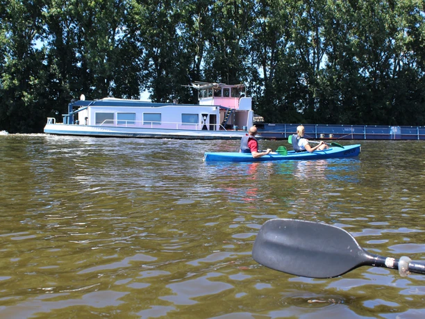 Kanu auf ruhigem Fluss, zwei Personen paddeln, im Hintergrund ein Schiff, umgeben von Bäumen.