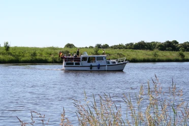Ein kleines Motorboot mit Personen fährt entlang eines ruhigen Flusses, umgeben von grüner Natur.