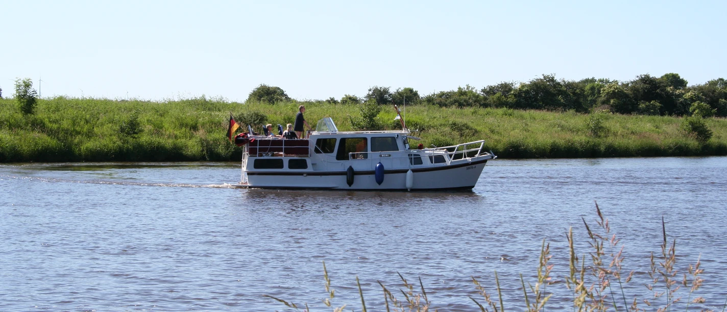 Ein kleines Motorboot mit Personen fährt entlang eines ruhigen Flusses, umgeben von grüner Natur.