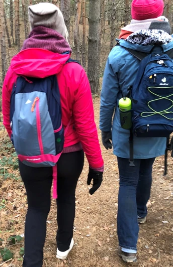Zwei Personen wandern in warmer Kleidung über einen schmalen Waldpfad, umgeben von hohen Bäumen.