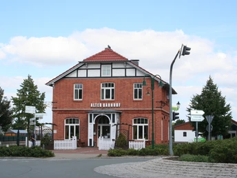 Ein historisches Backsteingebäude mit weißen Fenstern und Türen, umgeben von Bäumen und Straßenschildern.