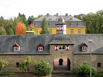 Schloss Heiligenhoven Historisches Schloss mit gelber Fassade, grauem Dach und buntem Herbstlaub im Hintergrund.