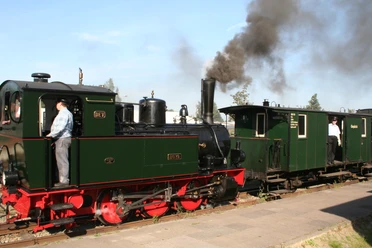 Steam locomotive pulls historic train on narrow-gauge railroad, two railroad enthusiasts stand on platforms.