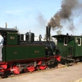 Steam locomotive pulls historic train on narrow-gauge railroad, two railroad enthusiasts stand on platforms.