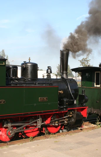 Steam locomotive pulls historic train on narrow-gauge railroad, two railroad enthusiasts stand on platforms.