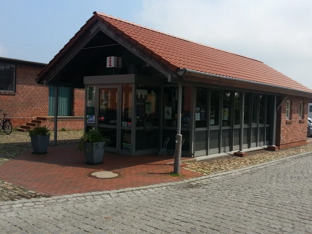 TourismusService Büro Einstöckiges Backsteingebäude mit roten Ziegeln, Glasfassade und Informationsschild an der Straße.