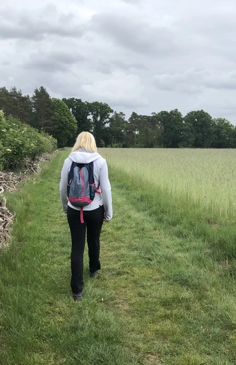 Eine Frau wandert auf einem grünen Pfad, umgeben von Feldern und Wäldern unter bewölktem Himmel.