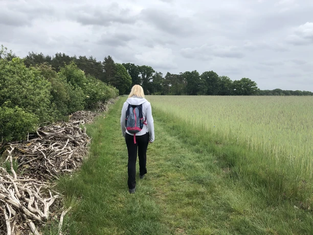 A woman walks along a green path surrounded by fields and woods under a cloudy sky.