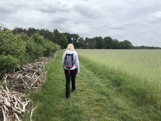 Wandern Eine Frau wandert auf einem grünen Pfad, umgeben von Feldern und Wäldern unter bewölktem Himmel.