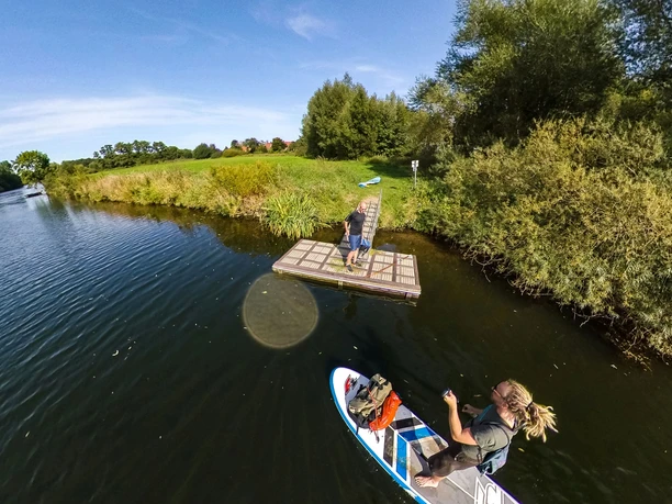 Rheine_SUP-Anleger_Thomas Mohn Fotografie Eine Person auf einem SUP-Board paddelt auf einem ruhigen Fluss neben einem Holzsteg.