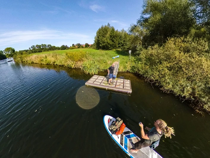 Rheine_SUP-Anleger_Thomas Mohn Fotografie Eine Person auf einem SUP-Board paddelt auf einem ruhigen Fluss neben einem Holzsteg.