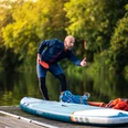 Mann in Sportbekleidung gestikulierend und lächelnd neben einem Stand-Up-Paddleboard auf einem Steg.
