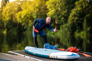 SUP and Hike - I like! Mann in Sportbekleidung gestikulierend und lächelnd neben einem Stand-Up-Paddleboard auf einem Steg.