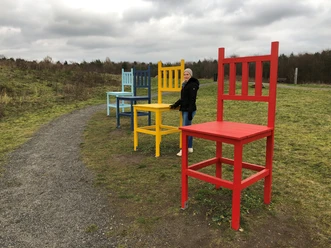 Colorful giant chairs on a dirt road, woman in winter clothes standing next to a red chair.