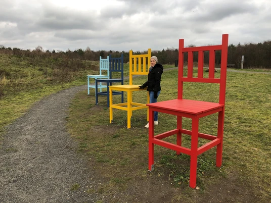Kräher Höhe Bunte Riesenstühle auf einem Feldweg, Frau in Winterkleidung steht neben einem roten Stuhl.