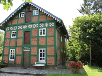 Ein zweistöckiges Fachwerkhaus mit rot-grünen Backsteinen und weißen Fenstern umgeben von Bäumen.