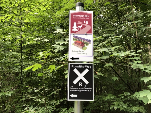 Beschilderung Beschilderung im Wald zeigt Promenadenweg und Roswitha-Weg, umgeben von dichtem grünen Laub.