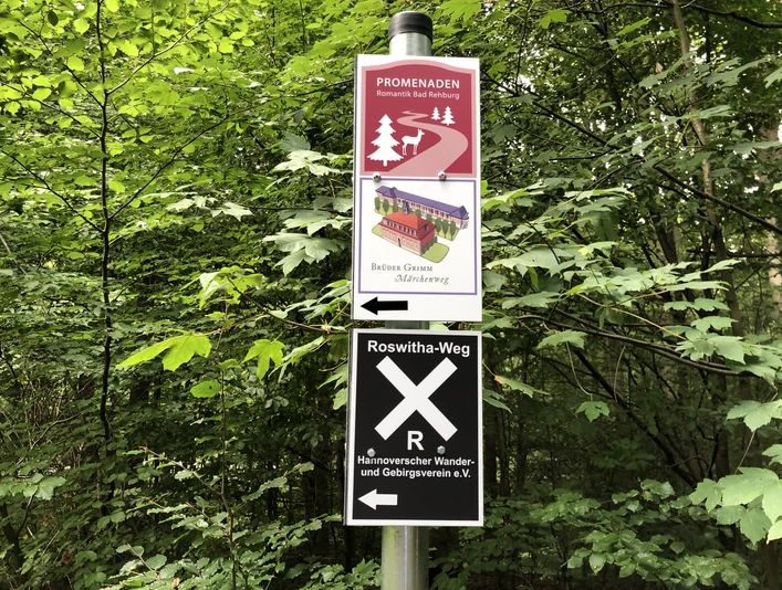 Beschilderung Beschilderung im Wald zeigt Promenadenweg und Roswitha-Weg, umgeben von dichtem grünen Laub.Signposts in the forest show the Promenadenweg and Roswitha-Weg, surrounded by dense green foliage.Skiltningen i skoven viser Promenadenweg og Roswitha-Weg, som er omgivet af tæt, grønt løv.Bewegwijzering in het bos toont Promenadenweg en Roswitha-Weg, omgeven door dicht groen gebladerte.