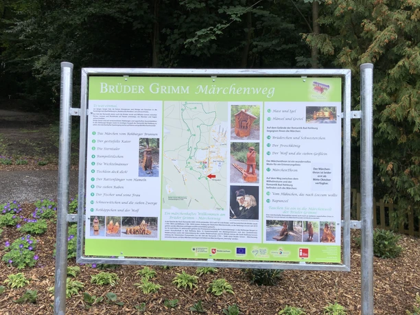 Infotafel Brüder Grimm-Märchenweg Information board of the Brothers Grimm Fairytale Trail in a green landscape, surrounded by plants and trees.