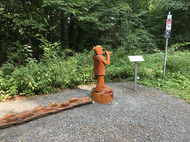 Houten figuur van een fluitspeler in het bos, omringd door sparren en een informatiebord aan de rand van het pad.