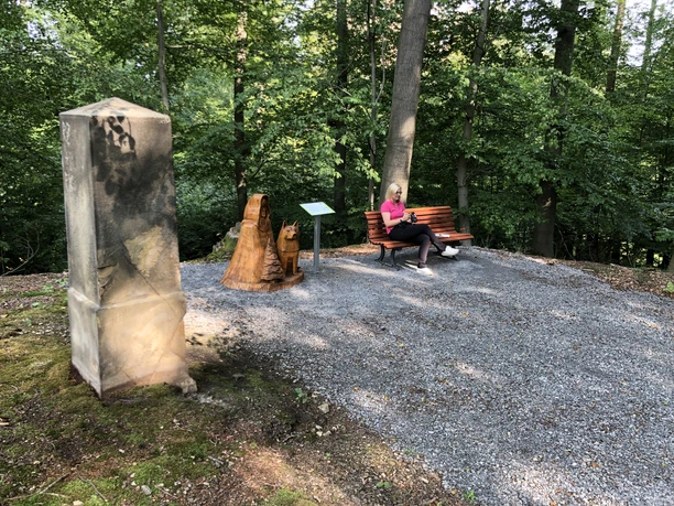 Een vrouw zit ontspannen op een bankje in een schaduwrijk bos, naast een houten beeld.
