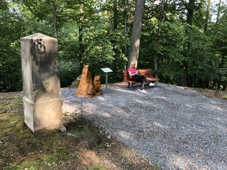 Promenaden Ernst August Höhe Eine Frau sitzt entspannt auf einer Bank in einem schattigen Wald, neben einer Holzskulptur.