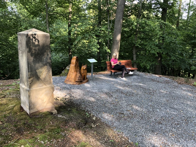 Promenaden Ernst August Höhe Eine Frau sitzt entspannt auf einer Bank in einem schattigen Wald, neben einer Holzskulptur.A woman sits relaxed on a bench in a shady forest, next to a wooden sculpture.En kvinde sidder afslappet på en bænk i en skyggefuld skov ved siden af en træskulptur.Een vrouw zit ontspannen op een bankje in een schaduwrijk bos, naast een houten beeld.