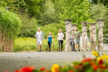Kurpark Nümbrecht <p>Fünf Personen wandern in sommerlicher Kleidung durch einen grünen Park mit antiken Säulen.</p>