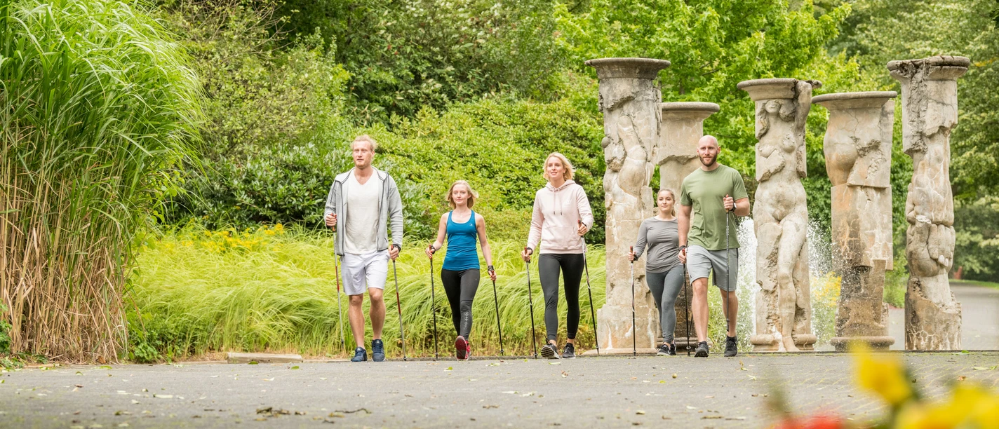 Kurpark Nümbrecht <p>Fünf Personen wandern in sommerlicher Kleidung durch einen grünen Park mit antiken Säulen.</p>
