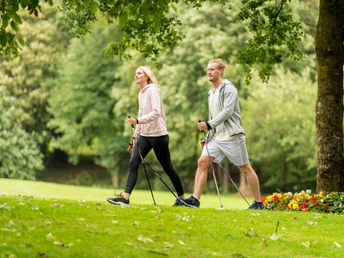 Fitness im Kurpark Nümbrecht Ein Paar beim Nordic Walking durch einen grünen Park, umgeben von Bäumen und Blumen.