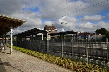 Bahnhof Lage Bahnhof mit überdachten Gleisen, Uhren, umliegenden Gebäuden, bepflanzter Fläche und blauem Himmel.