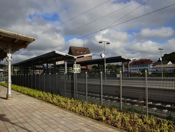 Bahnhof Lage Bahnhof mit überdachten Gleisen, Uhren, umliegenden Gebäuden, bepflanzter Fläche und blauem Himmel.