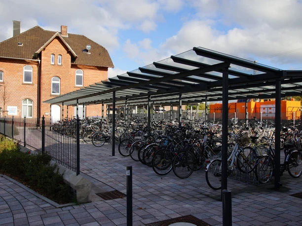 Bike & Ride Überdachtes Fahrradabstellplatz mit vielen geparkten Rädern vor einem historischen Backsteingebäude.