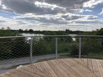 Eine hölzerne Aussichtsplattform mit Metallgeländer ermöglicht weiten Blick über grüne Landschaft und Gewässer.