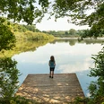 Herrenteich <p>Ein Kind steht auf einem Holzsteg, blickt über einen ruhigen See, umgeben von grüner Natur.</p>