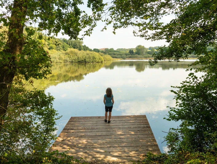 Herrenteich <p>Ein Kind steht auf einem Holzsteg, blickt über einen ruhigen See, umgeben von grüner Natur.</p>