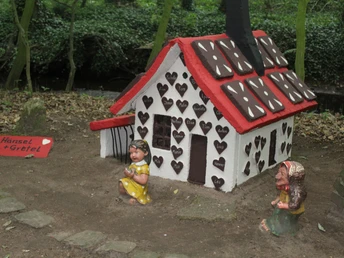 Lebkuchenhaus im Wald, umgeben von üppigem Grün, dargestellt mit Figuren von Hänsel und Gretel.