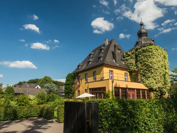 Burg Zweiffel Gelbes, historisches Haus mit Türmchen, umgeben von grüner Landschaft unter blauem Himmel.