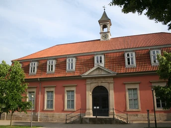 Längliches Gebäude mit rotem Ziegeldach, Uhrturm, symmetrischen Fenstern und einem steinernen Eingang.