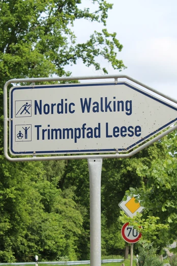Nordic Walking und Trimpfad Leese Hinweisschild weist auf Nordic Walking und Trimpfad Leese, umgeben von grünen Bäumen, nahe Landstraße.