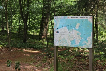 Wegwijzer in het bos met een kaart voor nordic walking en het Leese fitnessparcours, omgeven door bomen en gebladerte.