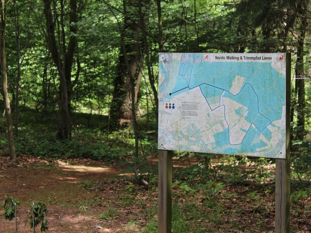 Wegwijzer in het bos met een kaart voor nordic walking en het Leese fitnessparcours, omgeven door bomen en gebladerte.