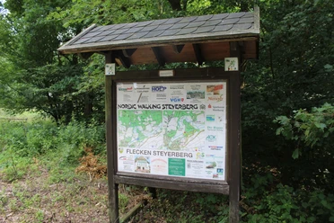 Houten bord met informatiekaart en sponsorlogo's in het bos, omringd door groen gebladerte en kreupelhout.