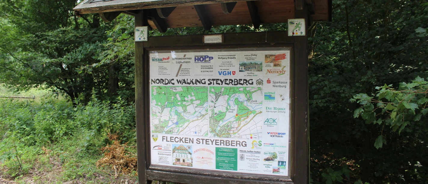 Houten bord met informatiekaart en sponsorlogo's in het bos, omringd door groen gebladerte en kreupelhout.