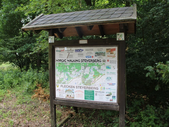 Holztafel mit Informationskarte und Sponsorenlogos im Wald, umgeben von grünem Blattwerk und Unterholz.