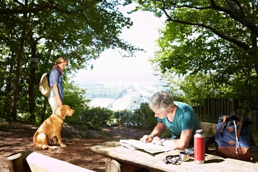 Wandern im Weserbergland Ein Wanderer rastet auf einer Lichtung im Weserbergland, blättert in einer Karte, sein Hund wartet.